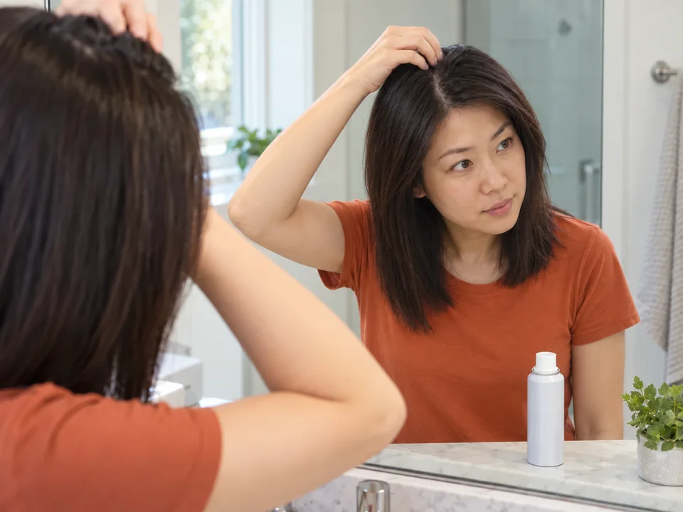 A woman parts her own dry hair at a mirror to inspect the roots.